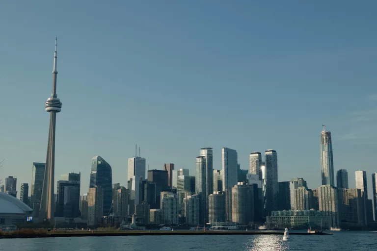 Panoramic Toronto skyline with the CN Tower on the left, seen across calm water with a small boat on a sunny day.
