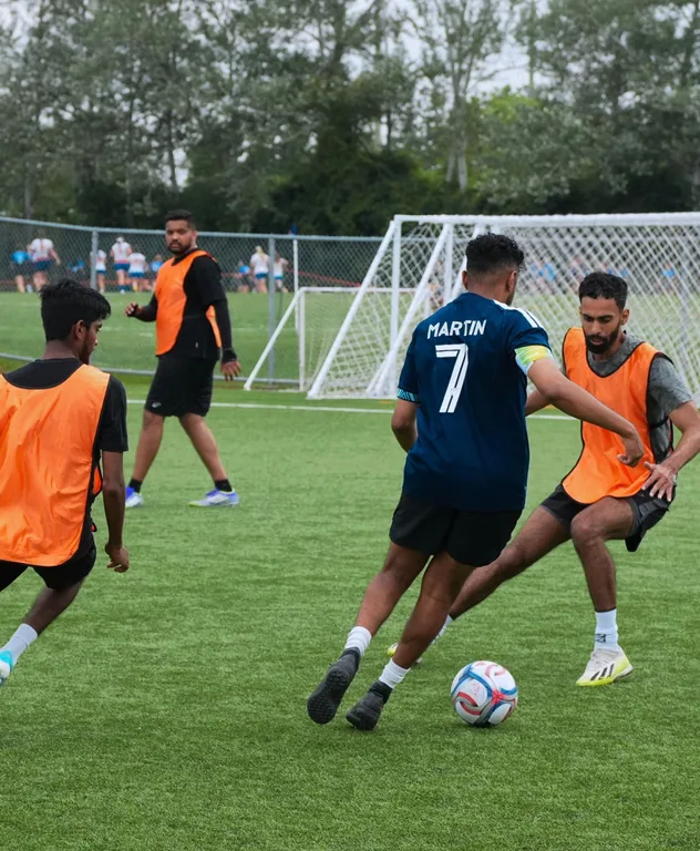 A soccer player in a navy jersey dribbling past a defender in an orange bib on an artificial turf field during training.