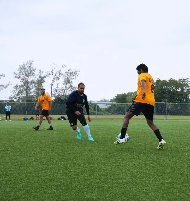 A soccer training run: a player in a yellow jersey moves with the ball while players in dark and orange jerseys close in around him on an outdoor turf field.