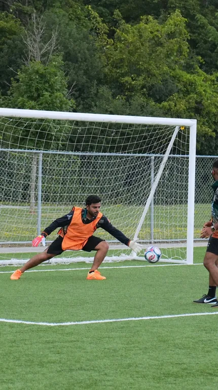 A soccer goalkeeper in an orange bib diving to his right to save a ball near the goal line on a turf field.