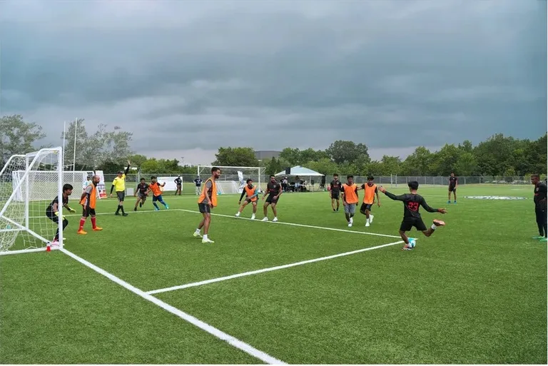 A soccer player in a black jersey striking the ball toward goal as defenders and a crouched goalkeeper react in the penalty area.